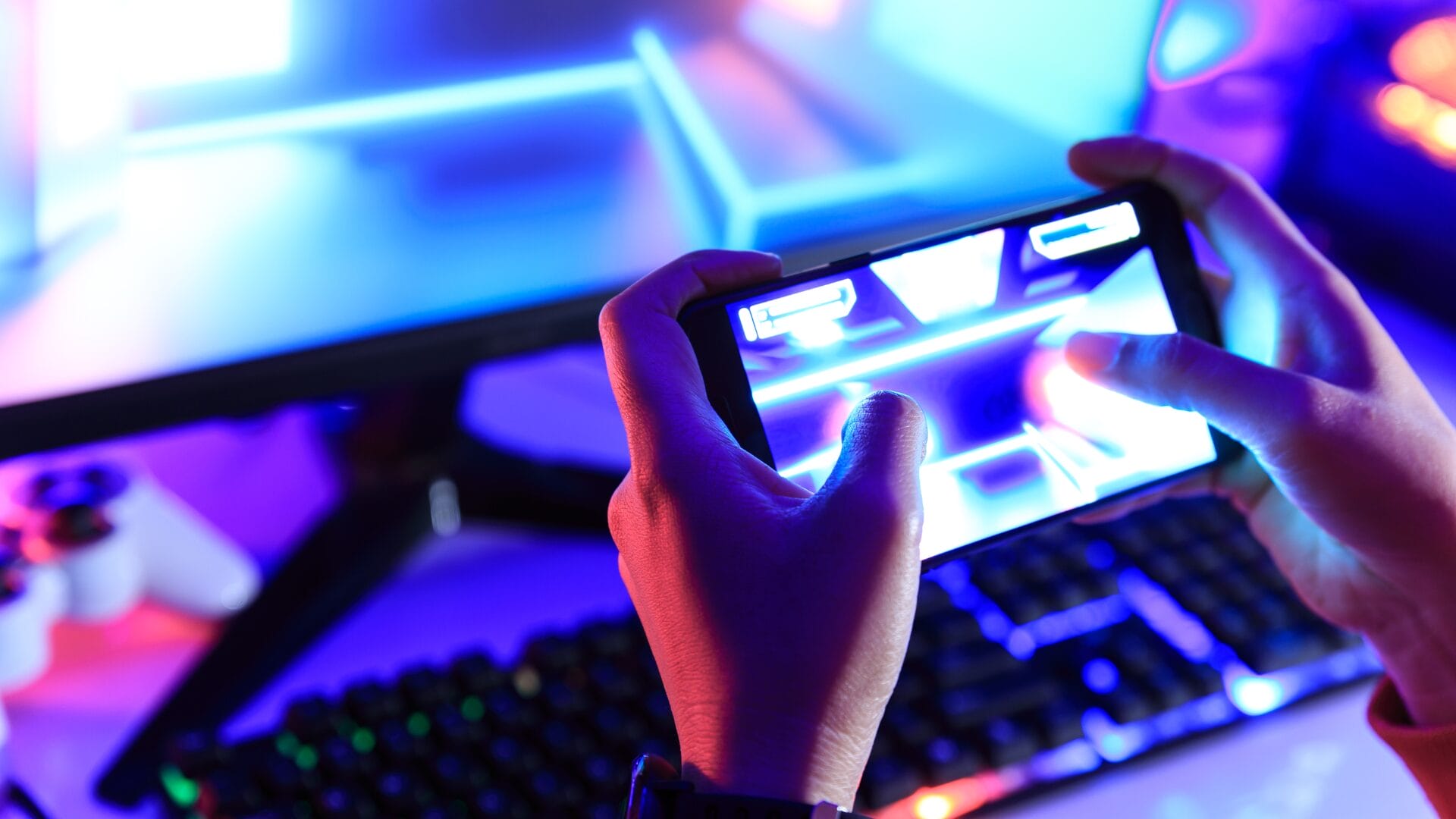 Hands of a person playing a mobile game with a vibrant gaming setup in the background, including a keyboard and a monitor.