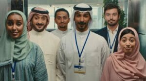 Group of six people in traditional Middle Eastern attire, including men in thobes and ghutras and women in hijabs, standing together in an elevator. The diverse group, featuring both men and women, appears slightly amused, reflecting the playful nature of McDonald’s localized ad campaign for the GCC region.