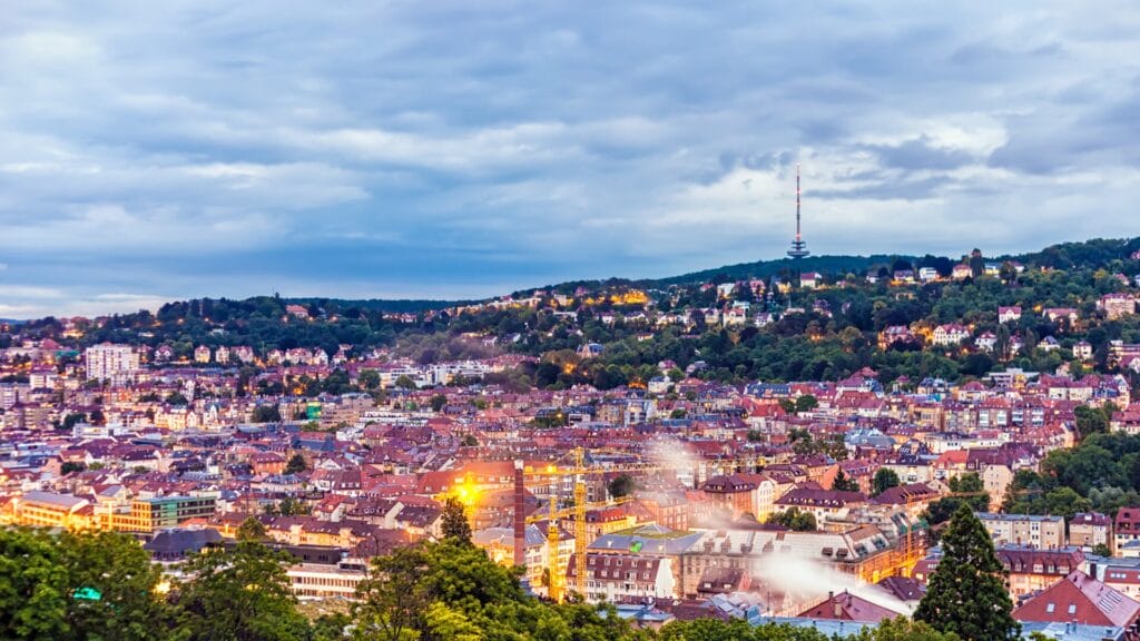 A panoramic view of Stuttgart, Germany at dusk, with houses and buildings nestled among green hills. The cityscape is softly illuminated, showcasing the area where the TCWorld Conference took place in 2024.
