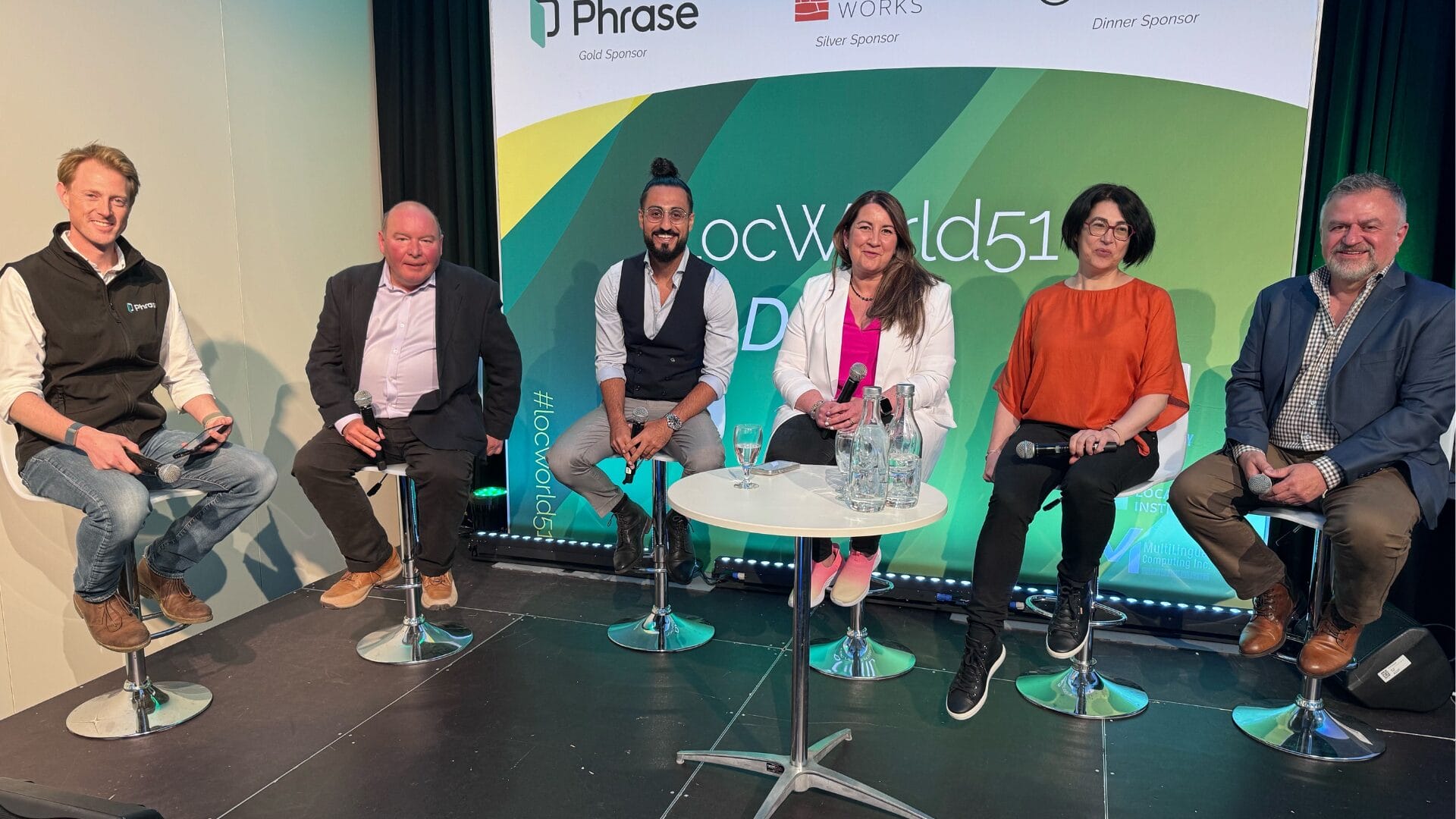 A panel of six people, including Georg Ell, CEO of Phrase, engaged in a discussion at LocWorld51. The panelists are seated on high stools in front of a backdrop displaying event sponsors. They are holding microphones and appear to be actively participating in the conversation.